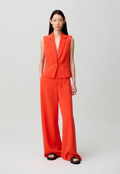 Donna in piedi che indossa un blazer senza maniche di un arancione acceso, pantaloni a gamba larga coordinati e sandali neri con suole spesse e fibbie.