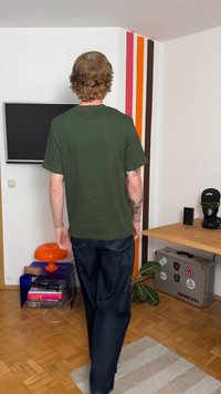 Olivově zelené tričko s krátkým rukávem a volným střihem, kombinované s volnými tmavými džíny. Světlá dřevěná podlaha a barevné stěnové akcenty v pozadí.