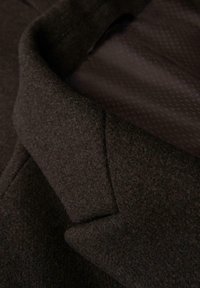 Hnědá vlněná látka zblízka, se strukturovaným povrchem. Límec má ostrý design, se hladkým vnitřním podšíváním s jemným vzorem.