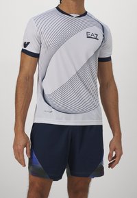 Muž oblečený v bílém sportovním tričku EA7 s zakřivenými černými pruhy a námořnických šortkách s modrými a zelenými akcenty, stojící před bílým pozadím.