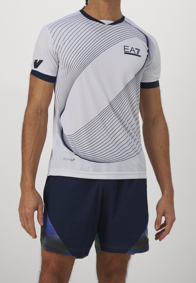 Muž oblečený v bílém sportovním tričku EA7 s zakřivenými černými pruhy a námořnických šortkách s modrými a zelenými akcenty, stojící před bílým pozadím.