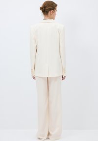 Krämfärgad pinstripe kostym med en skräddarsydd kavaj och vida byxor. Har knappr detaljer på ärmarna och ett sprund bak på kavajen.