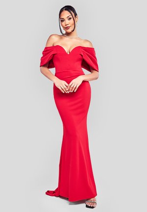 Femme en robe rouge ajustée à épaules dénudées avec des manches froncées, collier de perles et sandales à talons ornées de bijoux, se tenant devant un fond uni.