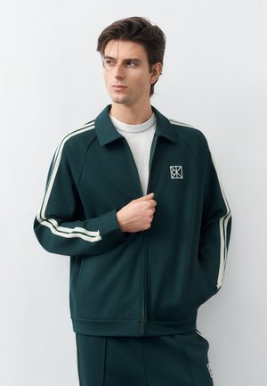 Mladý muž v tmavozelenej športovej bunde na zips s bielymi pruhmi na rukávoch a s rovnakými nohavicami, stojaci pred bielym pozadím.