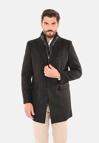 Uomo con barba che indossa un soprabito nero e pantaloni beige, che sistema il colletto del soprabito, in piedi su sfondo bianco.