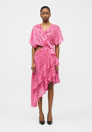 Žena stojaca v ružových asymetrických šatoch s volánmi a abstraktnými škvrnami, obutá v čiernych špicatých lodičkách na bielom pozadí.