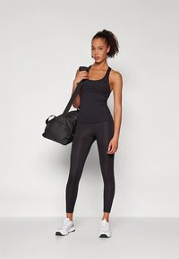 Vrouw in een zwarte sporttanktop en legging die een zwarte sporttas over haar schouder houdt, staande tegen een eenvoudige witte achtergrond.