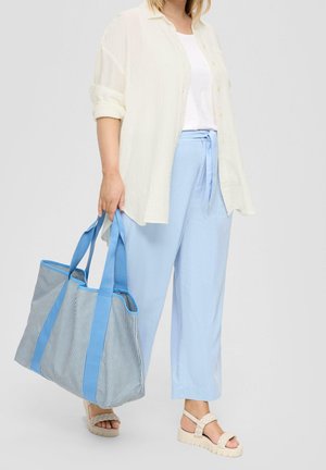 Person trägt ein weißes Hemd, hellblaue Hosen mit weitem Bein und cremefarbene Sandalen und hält eine große gestreifte Tragetasche mit blauen Griffen.