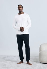 Λευκό πουκάμισο με μακριά μανίκια και μαύρο παντελόνι φόρμας. Το πουκάμισο έχει υφή, ενώ το παντελόνι διαθέτει ριμπ μανσέτες στους αστραγάλους. Το μοντέλο στέκεται ξυπόλητο πάνω σε ανοιχτό γκρι χαλί.