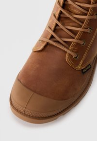 Bota de cuero marrón con punta de goma, ojales de metal y cierre de cordones. Superficie texturizada y suela de goma cosida. Etiqueta de marca visible.