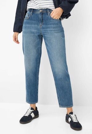 Persoon met een korte blauwe spijkerbroek, marineblauwe en witte sneakers, gestreept shirt en marineblauwe jas, staand met één hand in de zak.