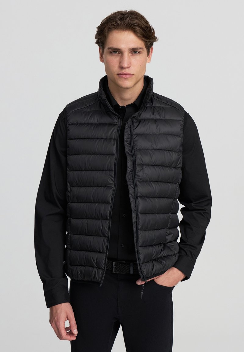 Gilet isolante nero con design trapuntato, dotato di colletto alto e chiusura con zip. Indossato sopra una camicia nera, crea un look a strati.