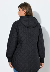 Giacca nera trapuntata con cappuccio, caratterizzata da un motivo a diamante e polsini a costine. La giacca ha un retro leggermente più lungo per una maggiore copertura.