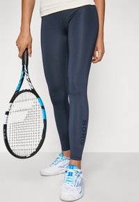 Tengerészkék teljesítmény leggings sima textúrával. Sötét színű "BORG" logó található az oldalán. Fehér cipővel és egy teniszütővel párosítva.