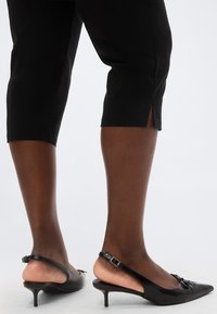 Άτομο με μαύρο παντελόνι έως τον αστράγαλο και μαύρες γόβες slingback με μυτερή μύτη και χαμηλό τακούνι kitten, στέκεται σε λευκό φόντο.