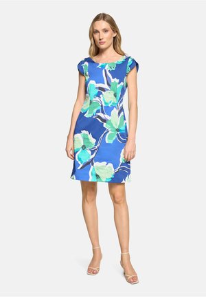 Frau steht vor einem schlichten Hintergrund und trägt ein blaues knielanges Kleid mit großem grün-weißem Blumenmuster sowie beige Riemchen-Absätze.