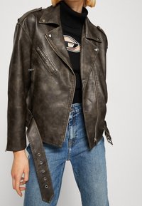 Veste de motard en simili cuir marron avec épaulettes, poches zippées et taille ceinturée, associée à un col roulé noir et un jean bleu.
