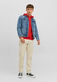 Chaqueta de mezclilla en azul claro, sudadera con capucha roja debajo, pantalones color crema y zapatillas negras con suela blanca, en pose de pie.