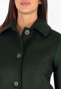 Cappotto di lana verde scuro con colletto, caratterizzato da grandi bottoni grigi e una superficie liscia, che evidenzia un design aderente.