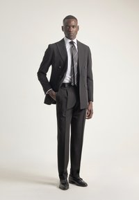 Hombre vestido con un traje negro a medida de doble botonadura, camisa blanca a rayas, corbata negra a rayas y zapatos de vestir negros, de pie frente a un fondo liso.