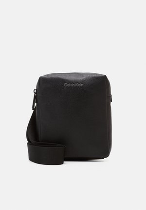 Czarna torebka na ramię Calvin Klein z fakturowaną skórzaną powierzchnią, regulowanym paskiem i srebrnym logo z przodu na jednolitym białym tle.