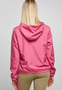 Blondynka w różowej kurtce z kapturem i beżowych spodniach, stojąca tyłem do kamery na jednolitym jasnym tle.