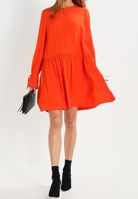 Punainen pitkähihainen mekko, jossa on valuva helma, kerrostettu vyötärö ja pehmeä kangas. Yhdistetty mustiin nilkkureihin ja käsilaukkuun.