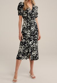 Černé květinové šaty s bílými květinami, přiléhavé korzetové tělo, objemné rukávy a délka po kolena. Obsahují přední rozparek a zavinovací design.