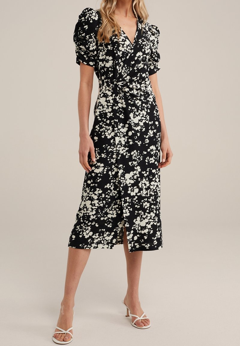 Černé květinové šaty s bílými květinami, přiléhavé korzetové tělo, objemné rukávy a délka po kolena. Obsahují přední rozparek a zavinovací design.