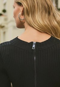 Black ribbed knit top features a rear zipper closure and a rounded neckline. The fabric texture is smooth with vertical lines.