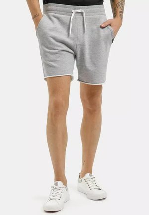 Osoba ubrana w jasnoszare szorty ze sznurkiem i białe trampki, stojąca z rękami w kieszeniach na białym tle.