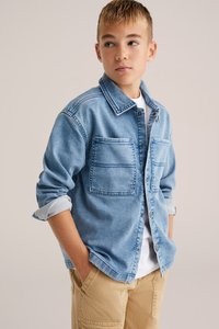 Šviesiai mėlyna džinsinė marškinė su dviem krūtinės kišenėmis, mygtukų užsegimu ir susuktais rankovėmis, derinami su smėlio spalvos cargo kelnėmis. Po jais uždėta paprasta balta marška.