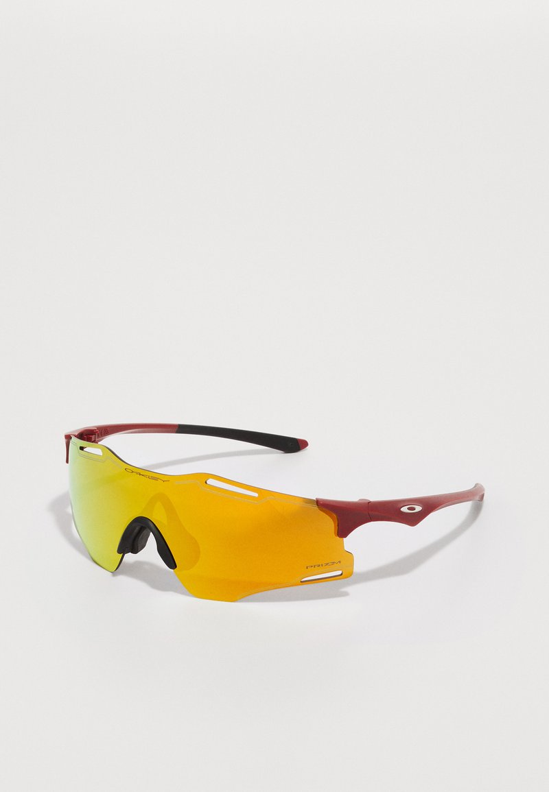 Óculos de sol desportivos com um design envolvente, lentes com tonalidade amarela, armação vermelha e pontas pretas. Apresentam recortes nas lentes para ventilação.