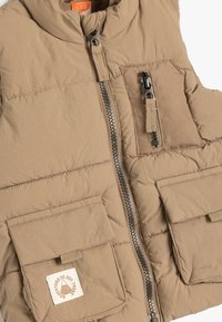 Chaleco acolchado beige con cuello alto, cremallera frontal y dos bolsillos delanteros. Presenta un parche con el logo en el bolsillo derecho. Tejido suave y texturizado.