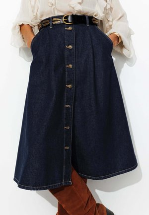 Jupe en denim en ligne A de couleur bleu foncé avec des boutons dorés sur le devant, dotée de poches latérales et d'une texture lisse. Ceinturée avec des accents en métal.