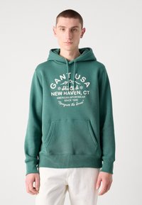 Hanorac verde cu buzunar tip cangur, decorat cu text grafic alb și un design montan. Textură moale, cu manșete și tiv zimțat. Pantaloni albi.