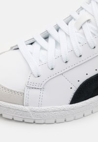 Gros plan sur une basket blanche avec des lacets blancs, un patch en daim noir, un embout gris et une semelle en caoutchouc texturée.