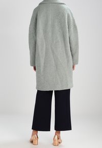 Cappotto sovradimensionato di un verde chiaro, texture accentuata, con colletto, visto da dietro, abbinato a pantaloni a gamba larga scuri e tacchi bronzo.