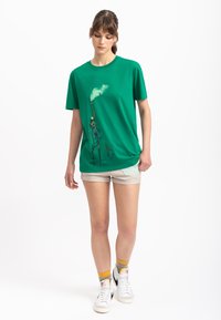 Groene katoenen T-shirt met een grafisch ontwerp van een figuur die een stok vasthoudt met rook. Korte mouwen, ronde hals, ontspannen pasvorm.