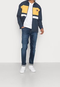 Marinblå jacka med gula och vita detaljer, med dragkedja framtill och logotyp-patch, i kombination med blå jeans och vita sneakers.