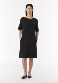 Robe noire à manches courtes avec un col rond, longueur genou, poches latérales et une texture lisse. Portée avec des chaussures noires.