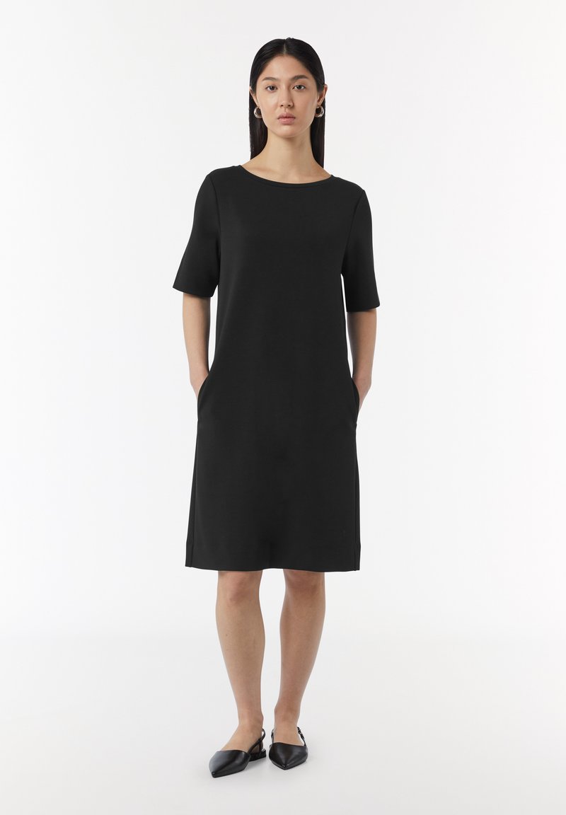 Robe noire à manches courtes avec un col rond, longueur genou, poches latérales et une texture lisse. Portée avec des chaussures noires.