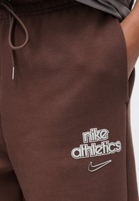 Καφέ παντελόνι τζόγκινγκ από απαλό ύφασμα, με λευκό λογότυπο "nike athletics" και ασημί σήμα Nike. Ελαστική μέση με κορδόνι.