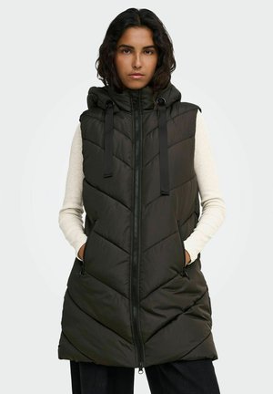 Frau trägt eine schwarze gesteppte ärmellose Kapuzenweste mit Frontreißverschluss und Taschen über einem hellen Langarmshirt und steht mit den Händen in den Taschen.