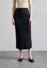 Zwarte, knielange rok van gladde stof, met een aansluitende taille en zijsplitten. Gecombineerd met zwarte ballerina's, tegen een neutrale achtergrond.