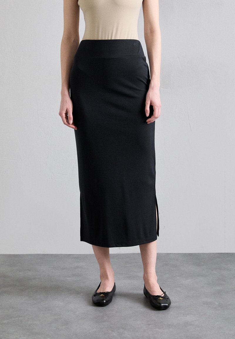 Zwarte, knielange rok van gladde stof, met een aansluitende taille en zijsplitten. Gecombineerd met zwarte ballerina's, tegen een neutrale achtergrond.