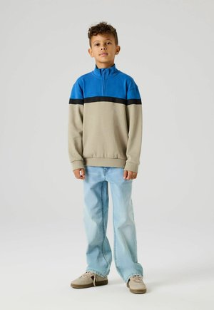 Poika seisoo päällään sininen ja beige vetoketjullinen collegepaita, vaaleansiniset farkut ja beiget tennarit yksinkertaisella vaalealla taustalla.