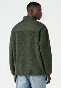 Veste en polaire verte avec un corps texturé et un panneau supérieur lisse. Elle présente un col haut et une coupe ample, portée avec un jean bleu clair.