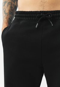 Schwarze Sweatpants mit einem elastischen Bund und einer Kordel, glatter Textur und seitlichen Taschen. Nahaufnahme von Nähdetails.