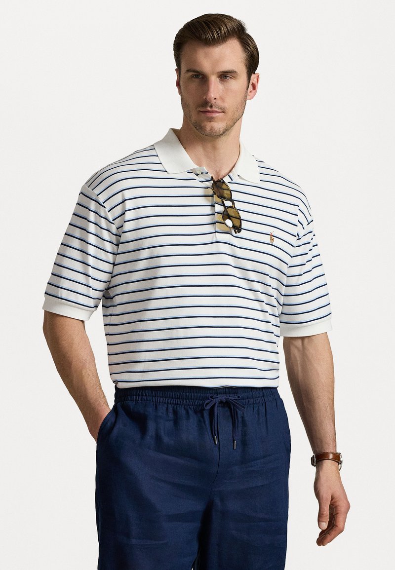 Camisa polo branca com riscas azul-marinho, de mangas curtas e colarinho. Usada com calções azul-marinho, óculos de sol pousados no colarinho.